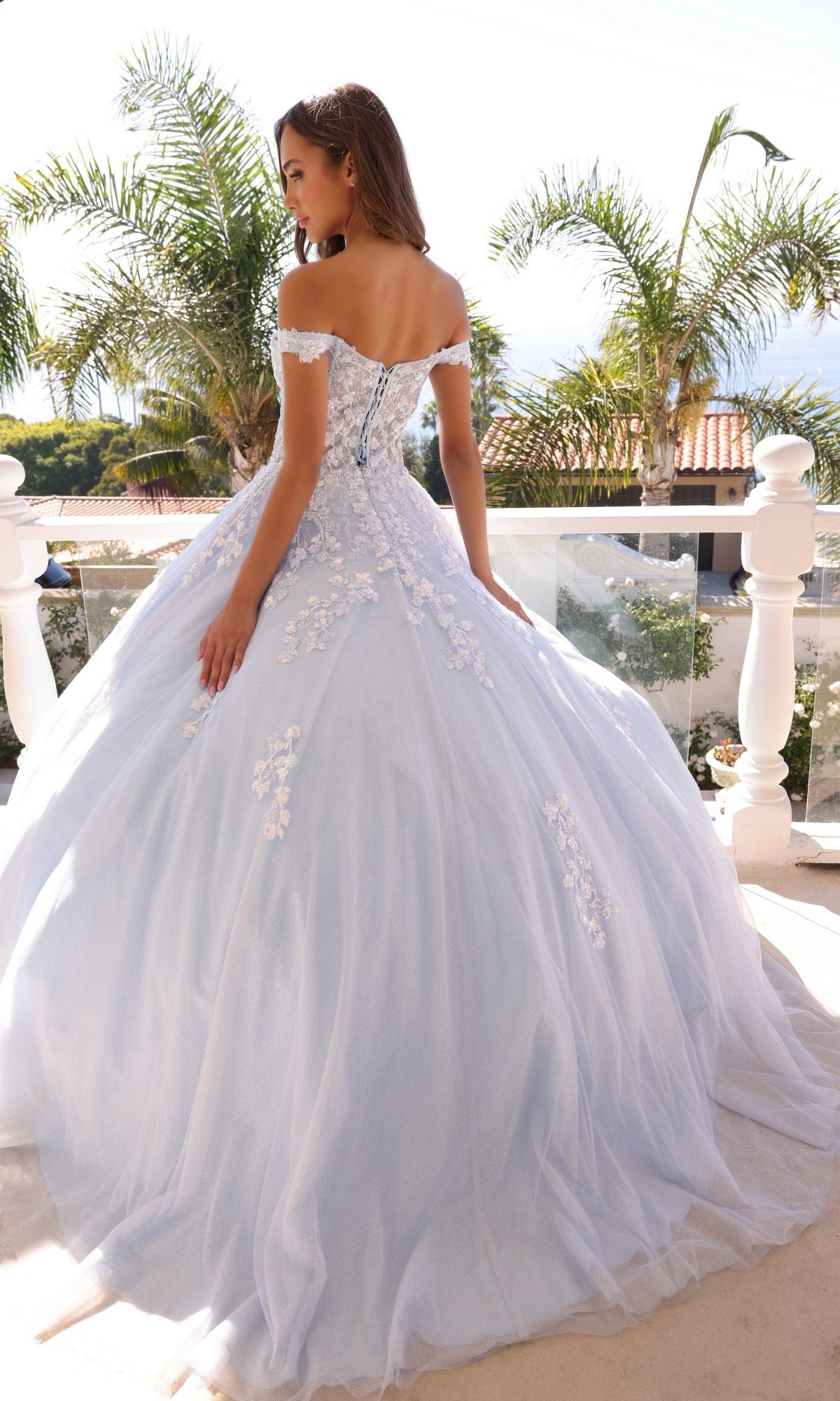 A woman stands on a balcony framed by palm trees, glowing in sunlight, wearing the Off-the-Shoulder Lace-Up Long Prom Ball Gown H1352 in pale blue with floral appliqué details.